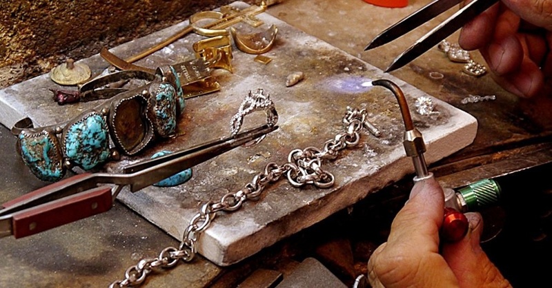 Jewelry tools and silver chains on a metal workbench with turquoise rings and a lock being opened