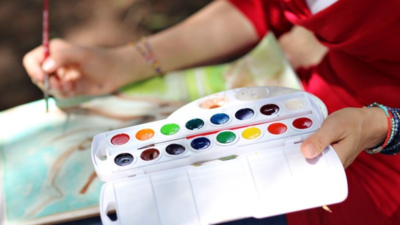 Hands holding a watercolor palette with multiple colored paint pans over an art sketch map