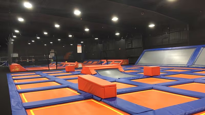 Indoor trampoline park arena with blue and orange padded trampolines and foam landing blocks under ceiling lights