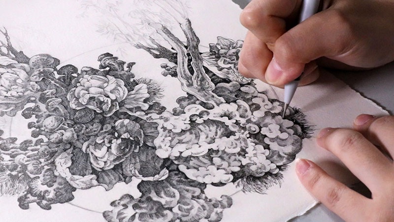 Hands drawing detailed black-and-white floral illustration with ink pen on white paper background