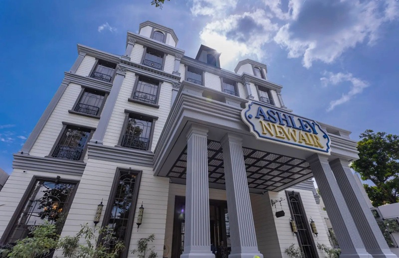 White neoclassical building with “Ashley” and “NEWLY” signage, tall columns, blue sky and clouds behind