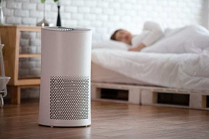 Air purifier with HEPA-style filter glowing in front of a bedroom with bed and person resting in background