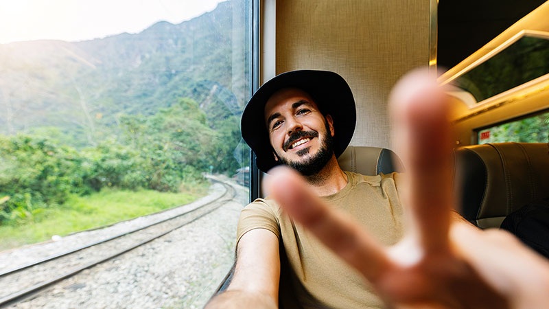 Man in a black hat smiling on a train, scenic mountain landscape through window and blurred hand in foreground