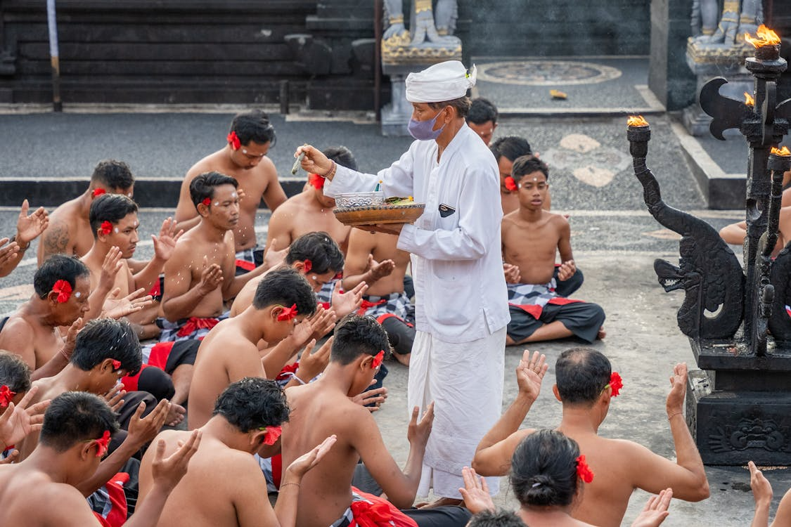 Bali temple ceremony as a priest in white offers blessings to shirtless men with flower petals in hand and raised palms