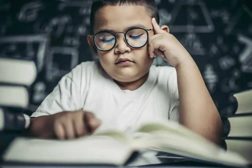 Child with glasses resting head on hand while reading an open book at a desk indoors