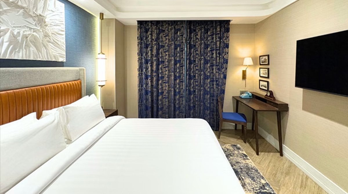 Modern hotel bedroom with king bed, patterned blue curtains, desk with lamp, and wall-mounted TV