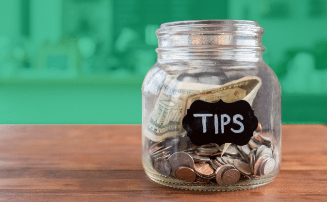 Glass jar labeled “TIPS” filled with coins on a wooden table against a blurred green background
