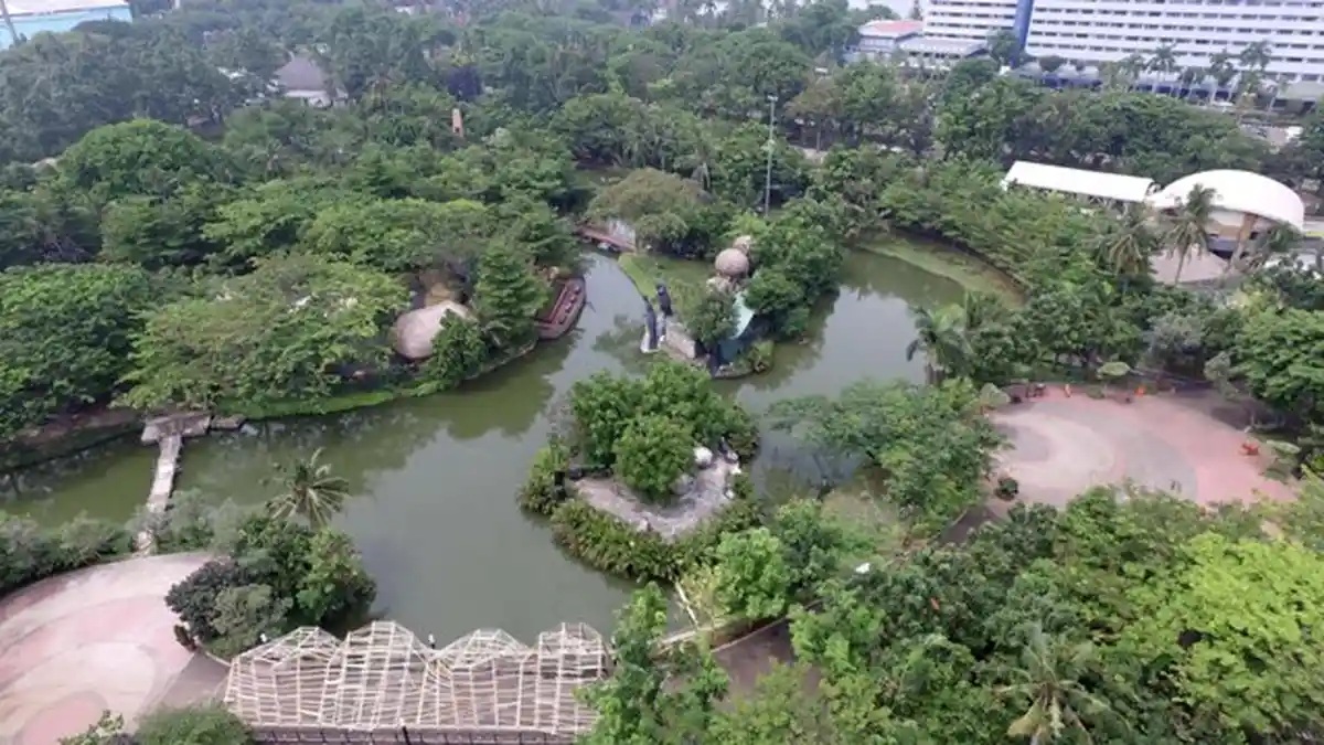 Aerial view of a lush river canal winding through dense greenery with a small bridge and buildings in the background