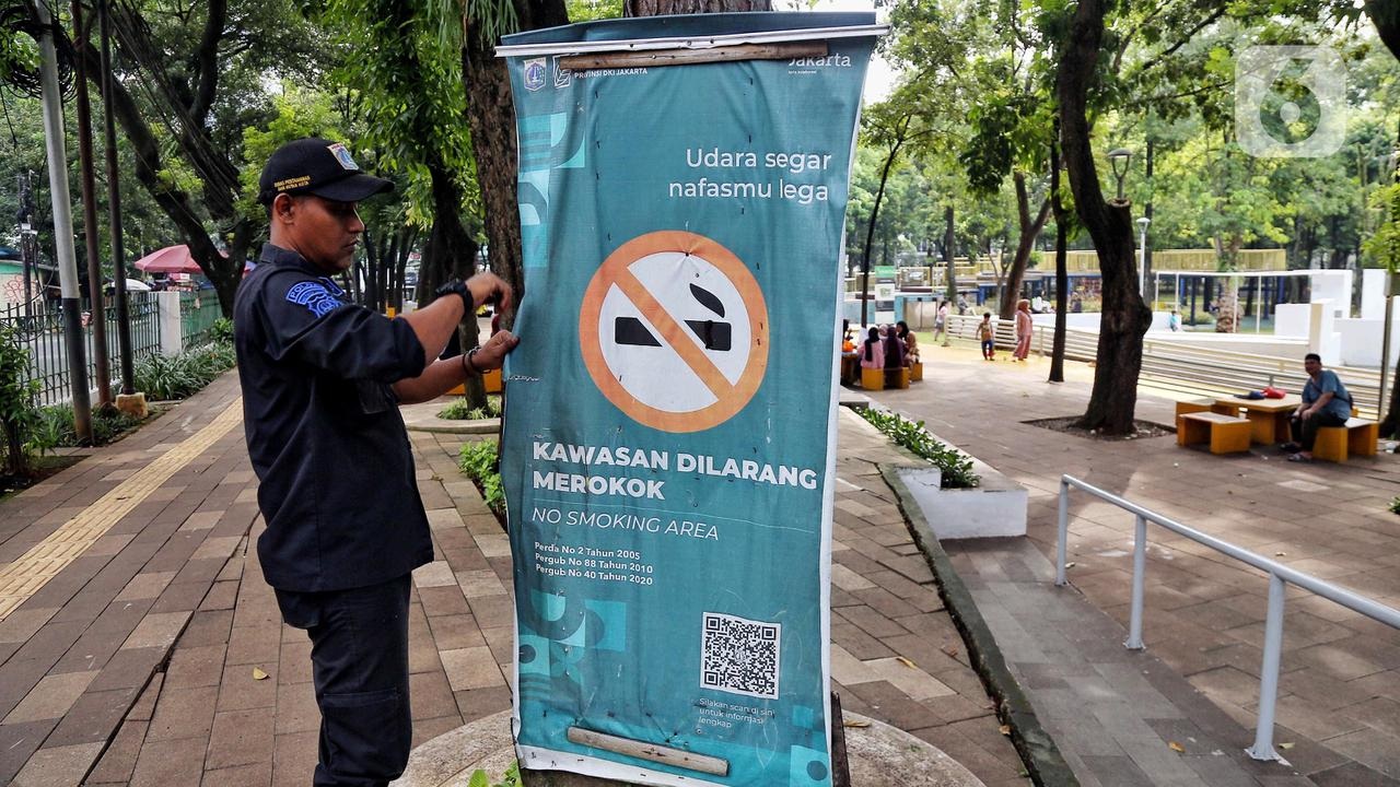 Street poster in Jakarta park reading “Kawasan Dilarang Merokok,” with no-smoking sign and a man standing beside it