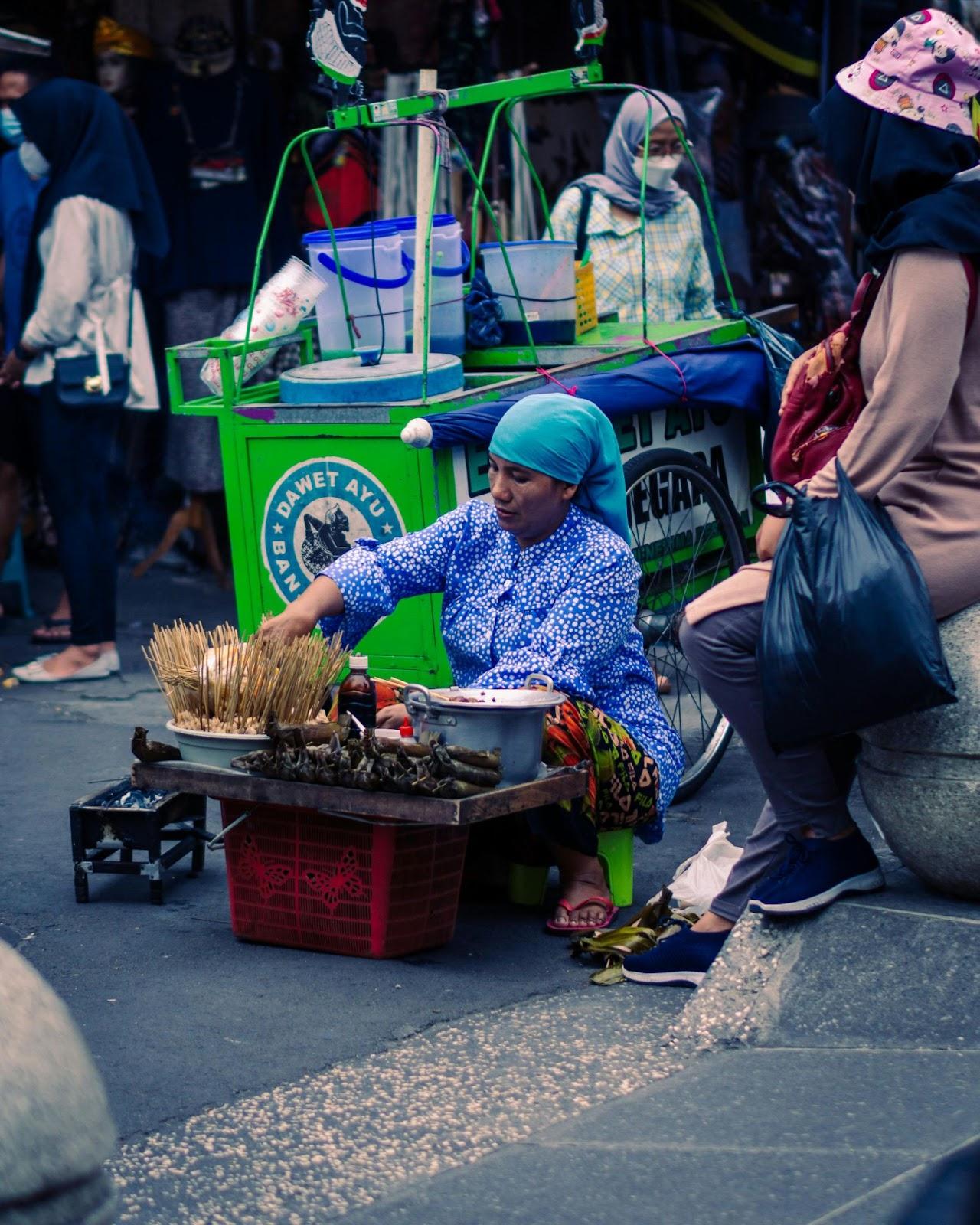 Street food vendor cooking with skewers at a green cart, seen with baskets, jars, and shoppers at night