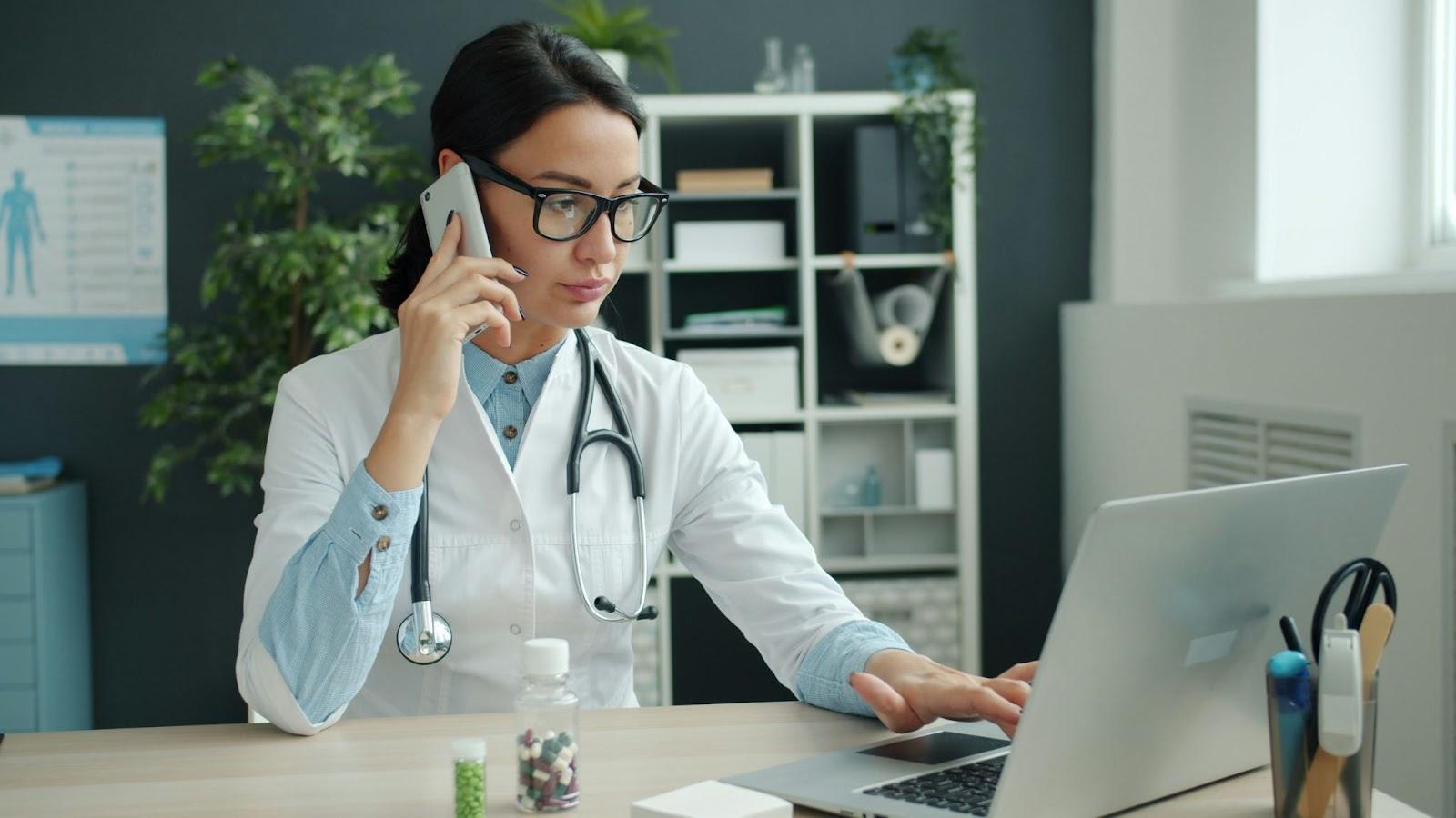 Doctor using laptop while speaking on phone in modern clinic office with stethoscope and paperwork