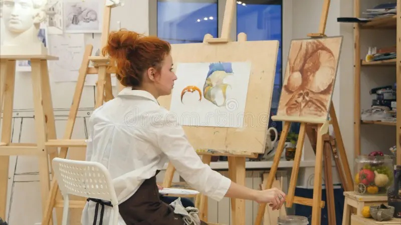 Artist painting on an easel in a studio, using a palette knife and canvas under bright lights