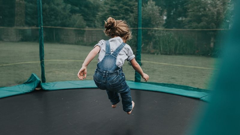 Toddler girl running on a backyard trampoline wearing denim overalls and a white shirt outdoors
