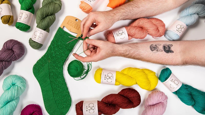 Hands weaving a green knit sock, surrounded by colorful yarn skeins on a white background