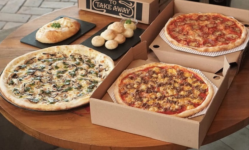 Assorted pizzas in cardboard boxes on a wooden table, plus garlic bread rolls and a takeaway sign