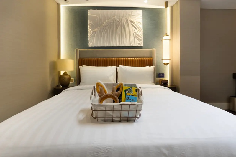 Modern hotel room bed with neatly made linens, bedside lamps, and a small welcome basket on the mattress