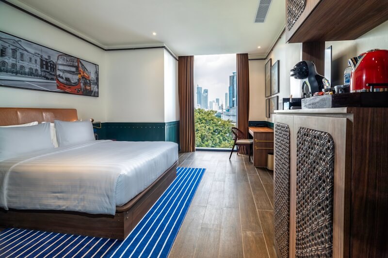 Modern hotel room with king-size bed, city skyline view through window, and wooden vanity with red accents