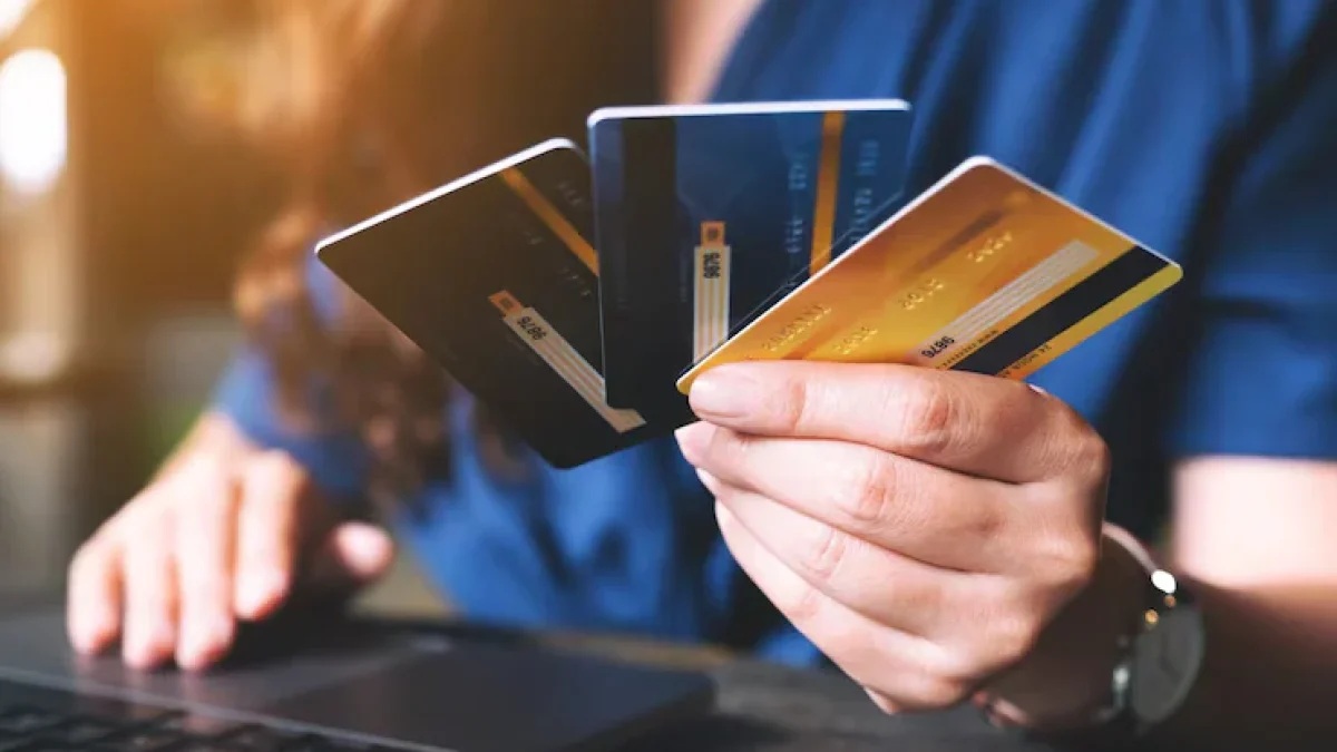 Hand holding credit cards and bank cards with a blurred person at a laptop on a table