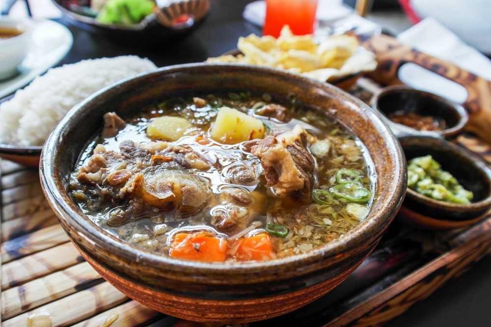 Hearty beef bone soup in a clay pot with carrots, potatoes, herbs, and scallions on a table