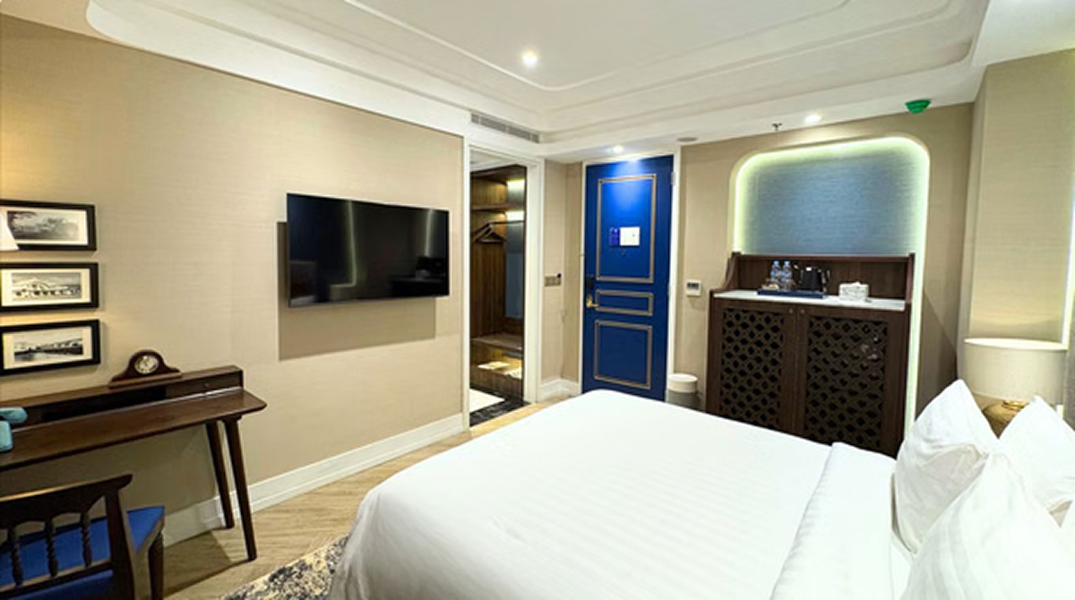 Modern hotel suite bedroom with white bed, blue door, TV wall, and reception-style desk with green LED trim