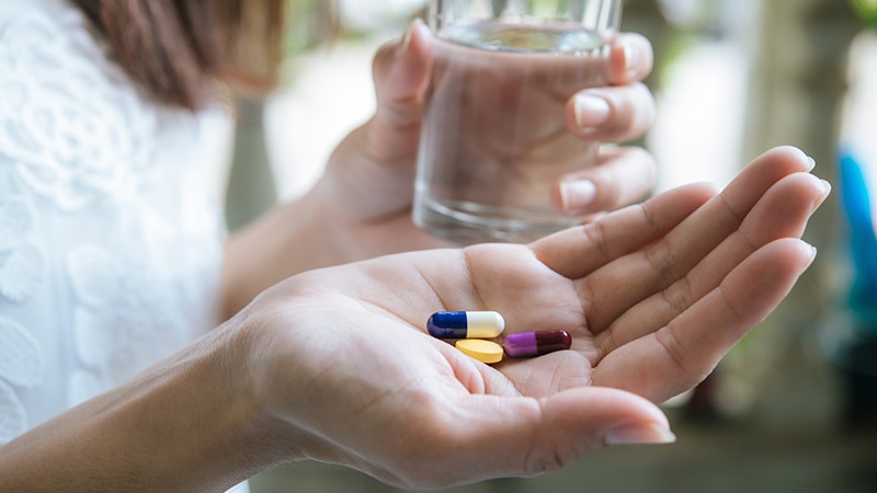 Hands holding pills and small capsules, with a person pouring water from a glass for medicine dosing