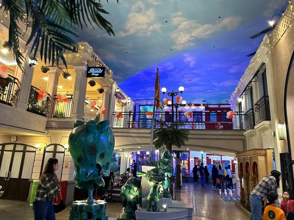 Outdoor shopping mall at dusk with festive lights, balloons, and shoppers walking between storefronts and arches