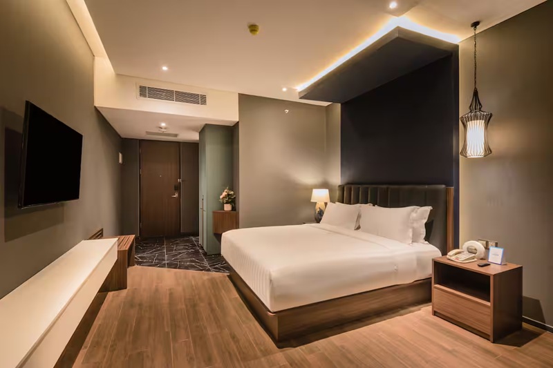 Modern hotel room with a neatly made bed, bedside tables, wall-mounted TV, and warm ceiling lights