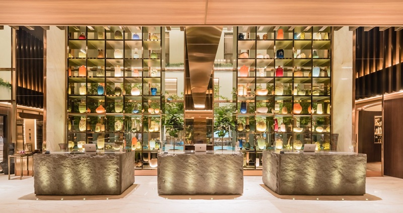 Luxury hotel lobby with rows of illuminated glass bottles and decorative topiary plants in gold display shelves