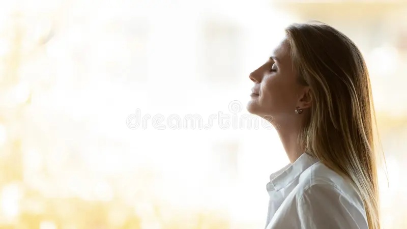 Woman in profile breathing outdoors in soft sunrise light, calm expression, copy space for wellness article