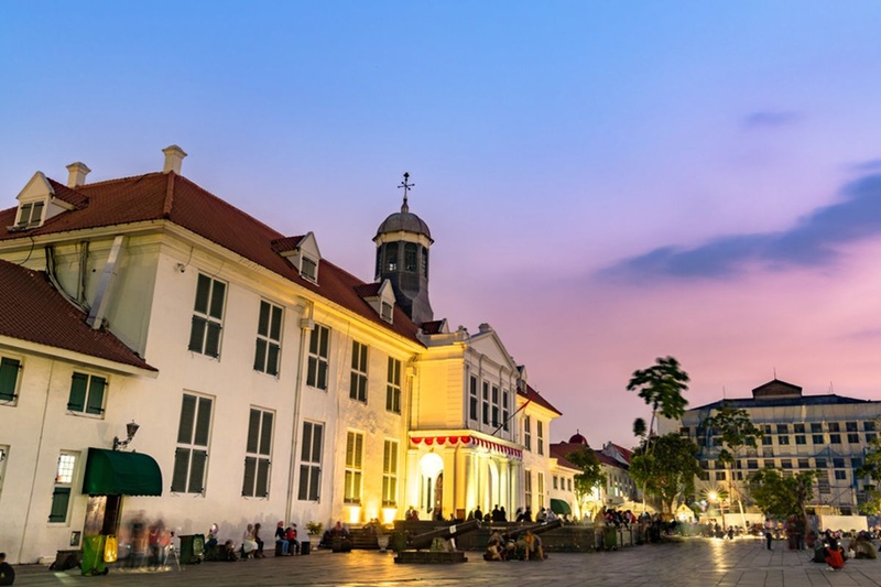 Historic campus building with clock tower at dusk, warm lights and people gathering in a courtyard plaza