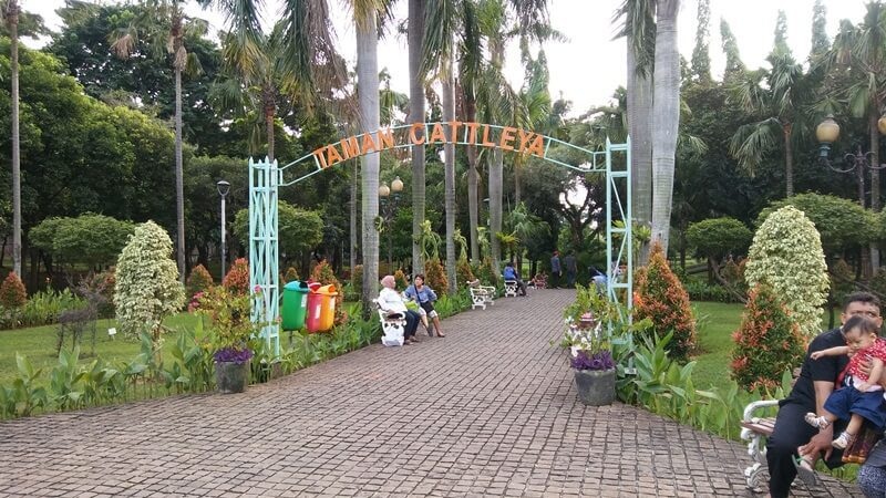 Taman Cattleya garden entrance with arch sign, paved walkway, potted plants and palm trees in the background