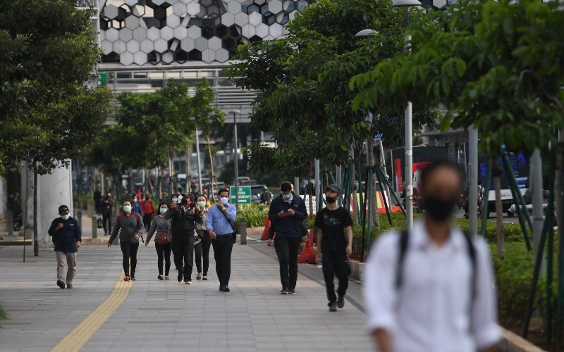 People walking on a city sidewalk wearing face masks, trees lining the walkway and storefronts in background