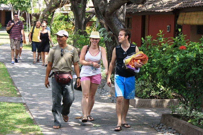 Tourists walking along a tropical garden path, one person carrying colorful towels, warm sunny day scenery