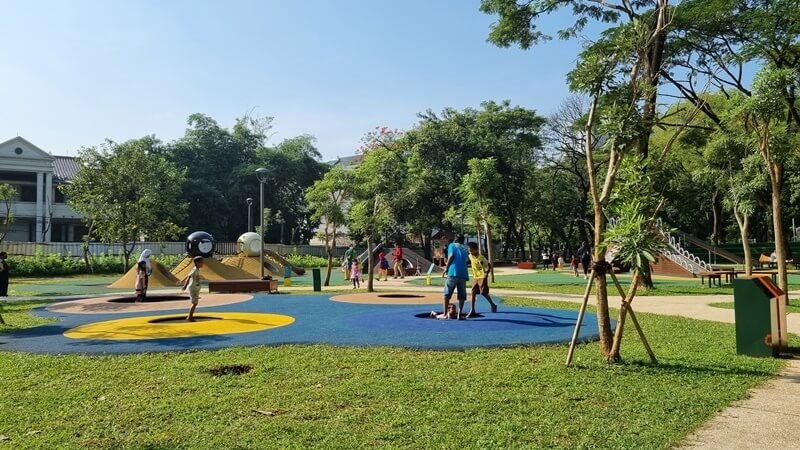 Sunny outdoor playground with kids walking on colorful rubber flooring and trees along pathways