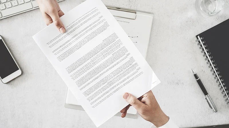 Hands holding a printed document with text on a desk with laptop, notebook, and pen nearby