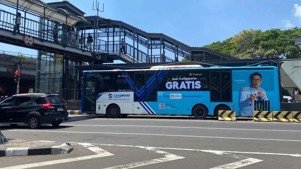 Blue public transit bus with “GRATIS” advertisement passing under an overpass on a city street, with parked cars nearby