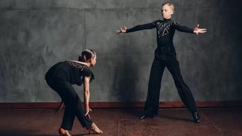 Dancer in black performing a stretching move on one leg while another dancer stands with arms out against gray wall