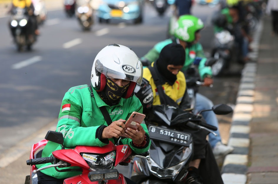 Motorcyclists in green racing jackets waiting on a busy street, helmeted rider checking a smartphone