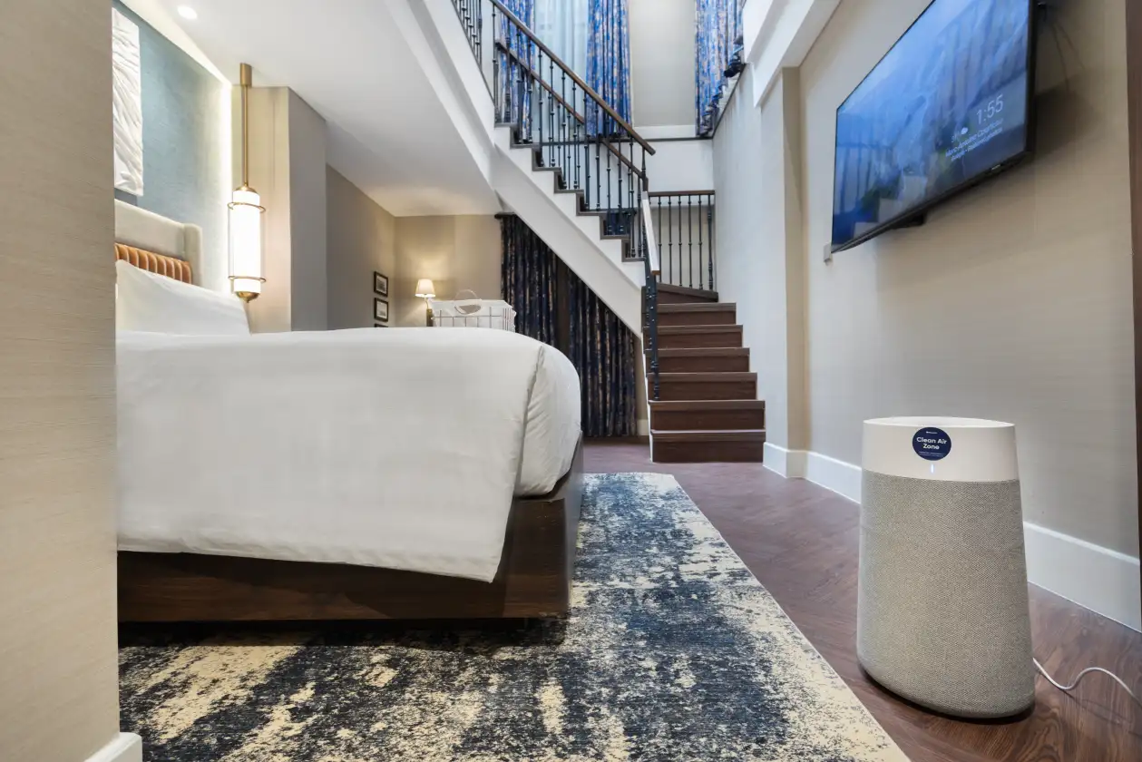 Clean Air Zone smart air purifier beside bed in modern bedroom with staircase, wall TV, and patterned rug