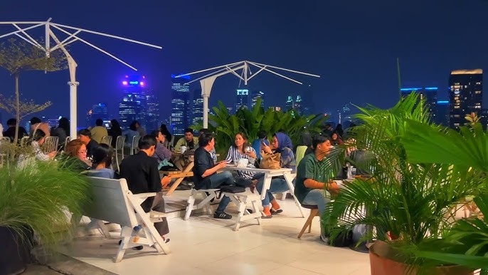 Outdoor rooftop dining area with guests seated at tables overlooking a modern city skyline at night