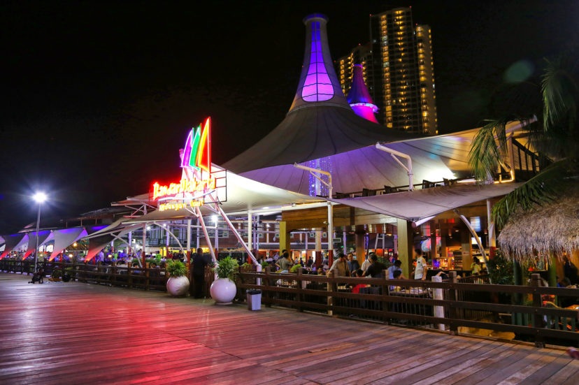 Night view of a waterfront restaurant patio with colorful lights, neon sign, and illuminated resort building with towers