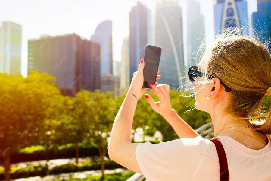 Woman taking a selfie with a smartphone, sunny city skyline background with trees and skyscrapers