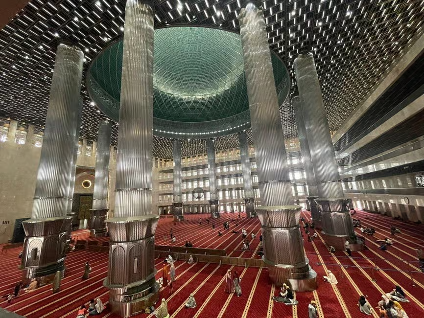 Interior of a grand mosque with towering columns, red carpet floors, and a large green domed ceiling