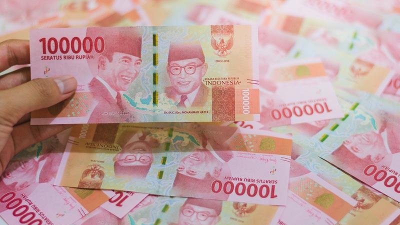 Hand holding Indonesian 100,000 rupiah banknote among scattered 10,000 and 100,000 rupiah bills