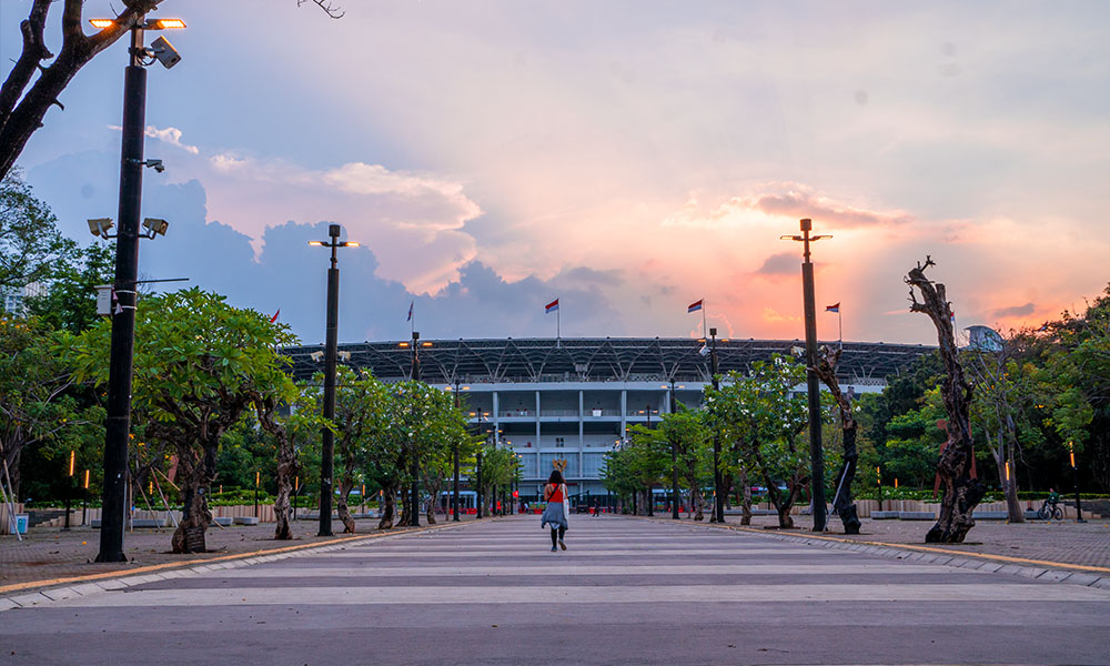 Outdoor view of a stadium with flagpoles and trees at sunset, with a person walking in the foreground