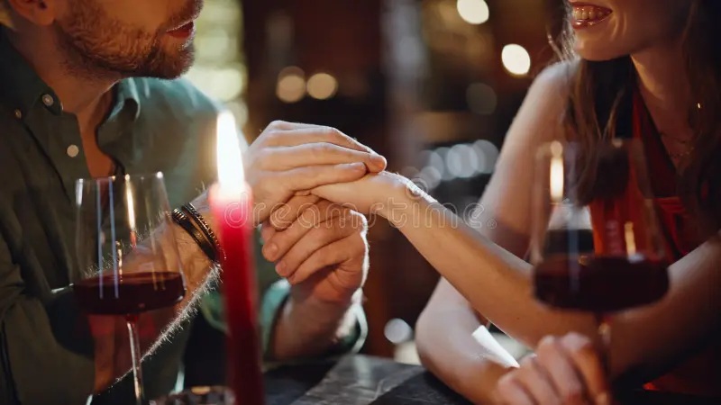 couple clinking hands while dining at a restaurant table, warm candlelight and wine glasses nearby