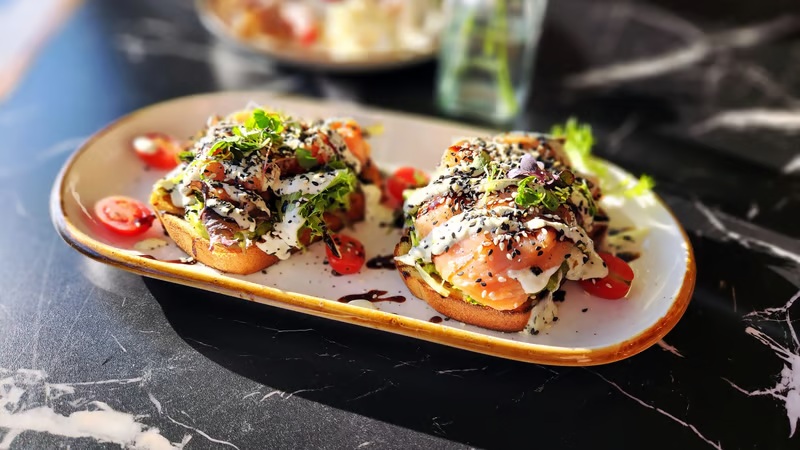 Chicken toast with poached eggs, herbs, sesame seeds, and sliced cherry tomatoes on a wooden platter