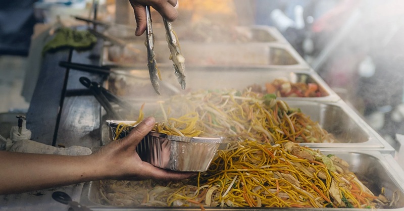 Street food vendor tossing spaghetti noodles on a large metal pan grill with tongs and serving tray