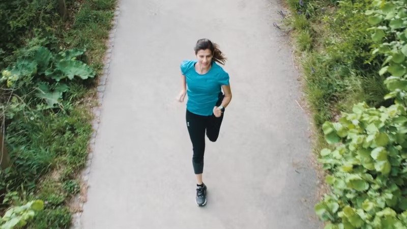 Woman jogging on a rural path surrounded by green bushes and trees, wearing a blue shirt and black leggings