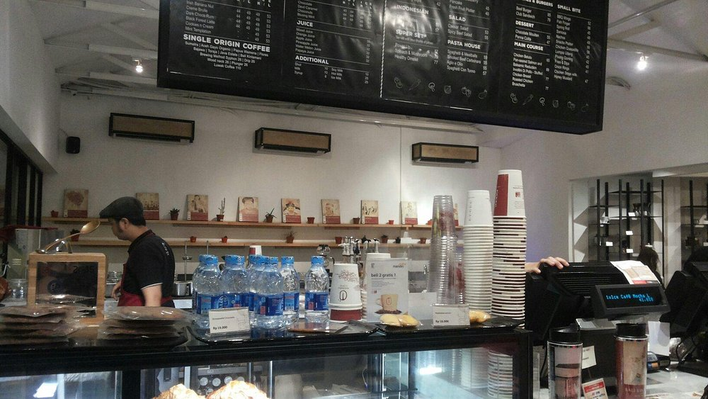 Coffee shop counter with barista, menu board, stacked cups, and beverage dispensers in Indonesia